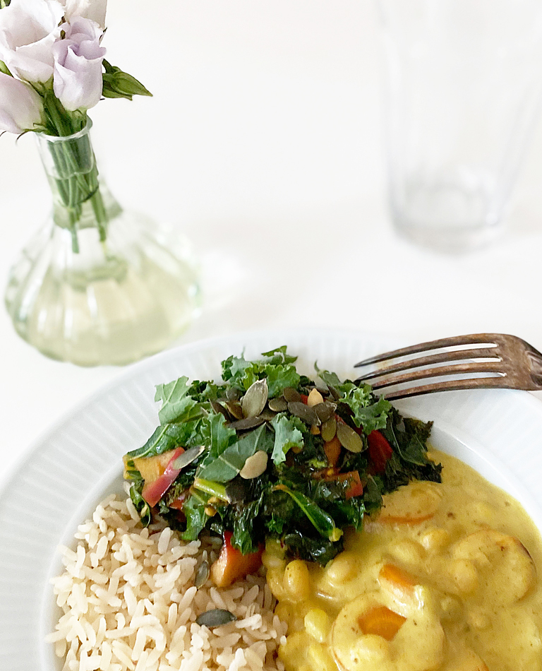 Ett fat med vegansk currygryta, ris och lättfräst grönkål står bredvid en vas med blommor.